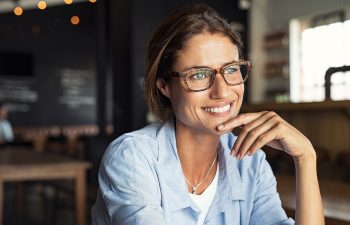 A woman wearing glasses and a blue shirt smiles, sitting in a dimly lit cafe., 