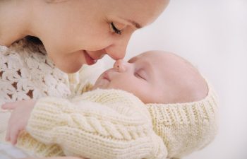 A woman gently touches her nose to a sleeping baby, both wearing knitted cream clothing., 