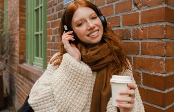 A person with red hair is wearing headphones and a scarf while holding a coffee cup, standing outside near a brick wall., 