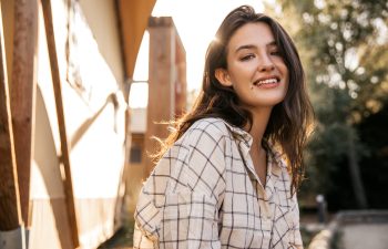 Young woman with long brown hair wearing a plaid shirt smiles outdoors in sunlight, with a blurred background of trees and buildings., 