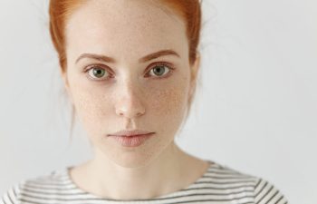 A young woman with red hair, green eyes, and freckles, wearing a white and black striped shirt, looks directly at the camera against a plain light background., 