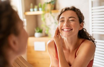 Woman smiling and applying skincare cream to her cheek while looking in the mirror in a bright bathroom with plants in the background.